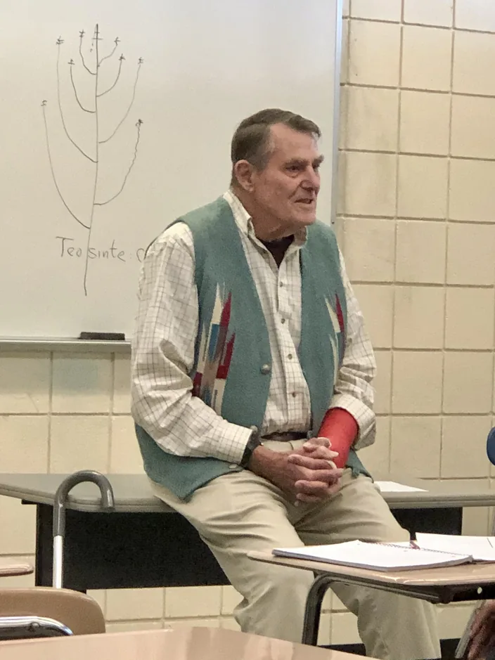 Garrison Wilkes seated on a desk, mid-lecture, with a diagram of a teosinte tassel drawn on the whiteboard behind him.